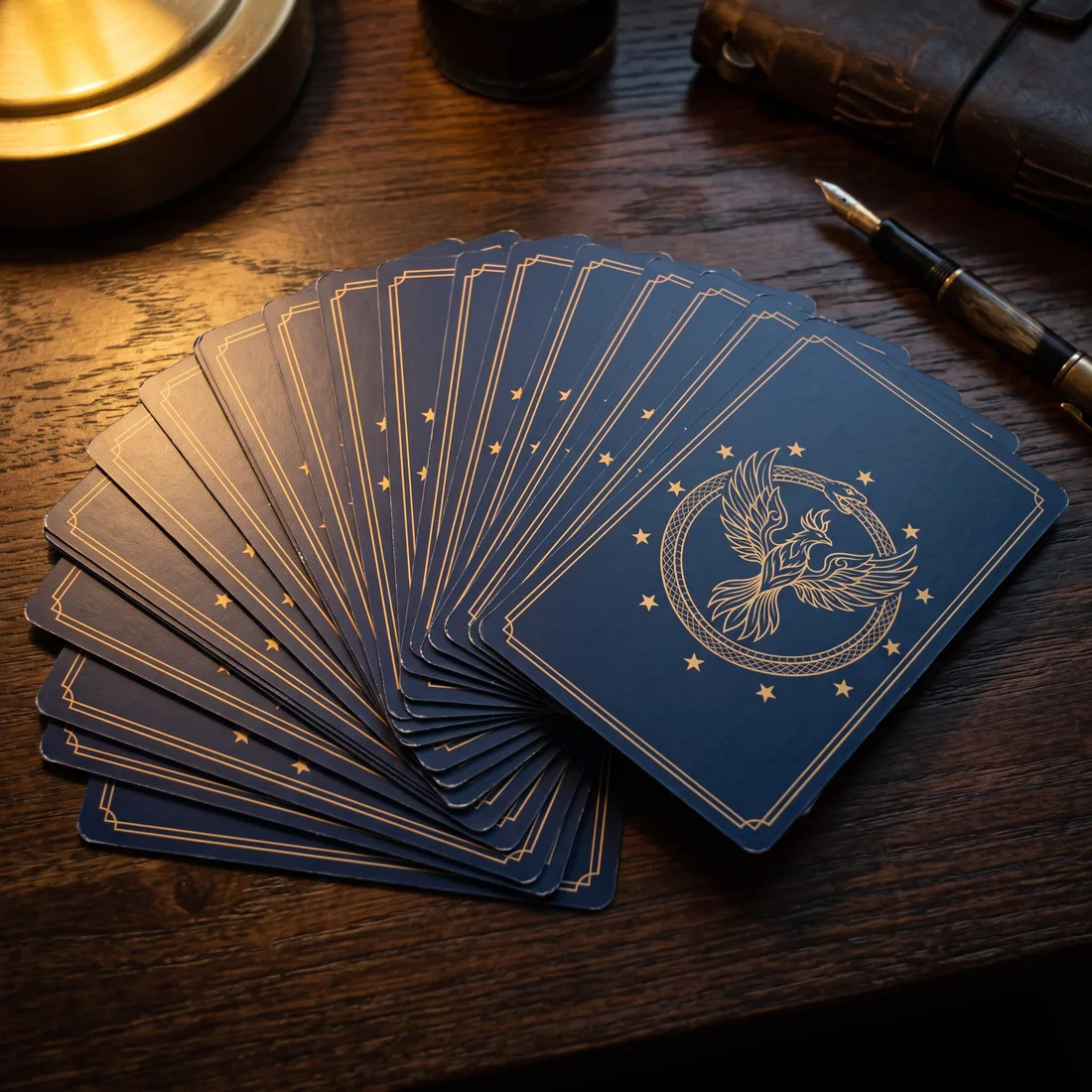 Les cartes du Codex imprimées et découpées à la main, étalées en éventail sur un bureau, face cachée : le phénix dans l'ouroboros, à l'or sur l'indigo. Un stylo plume repose à côté.